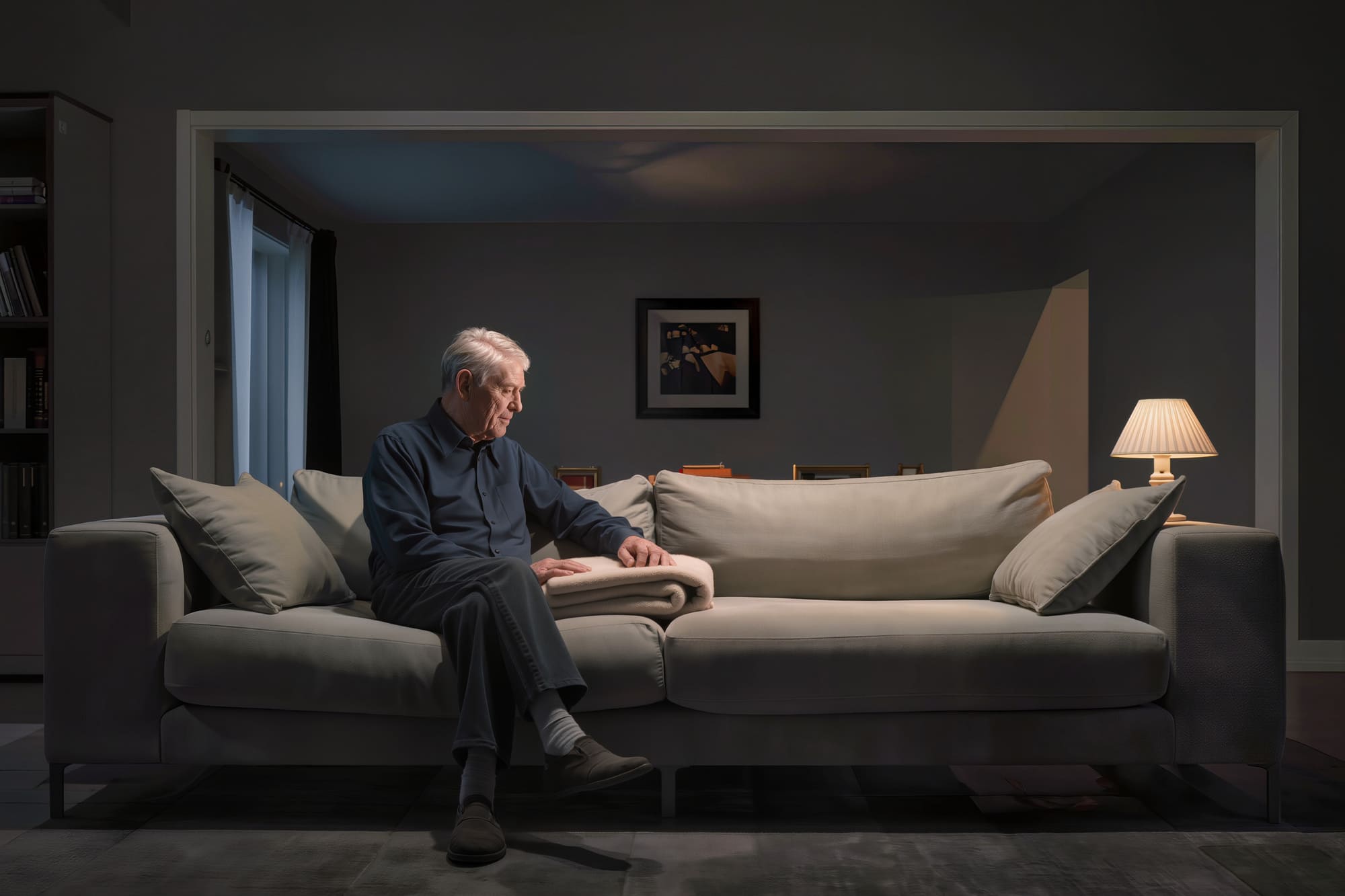 Homme âgé assis seul sur un canapé dans un salon sombre, illustrant la solitude et l’isolement.